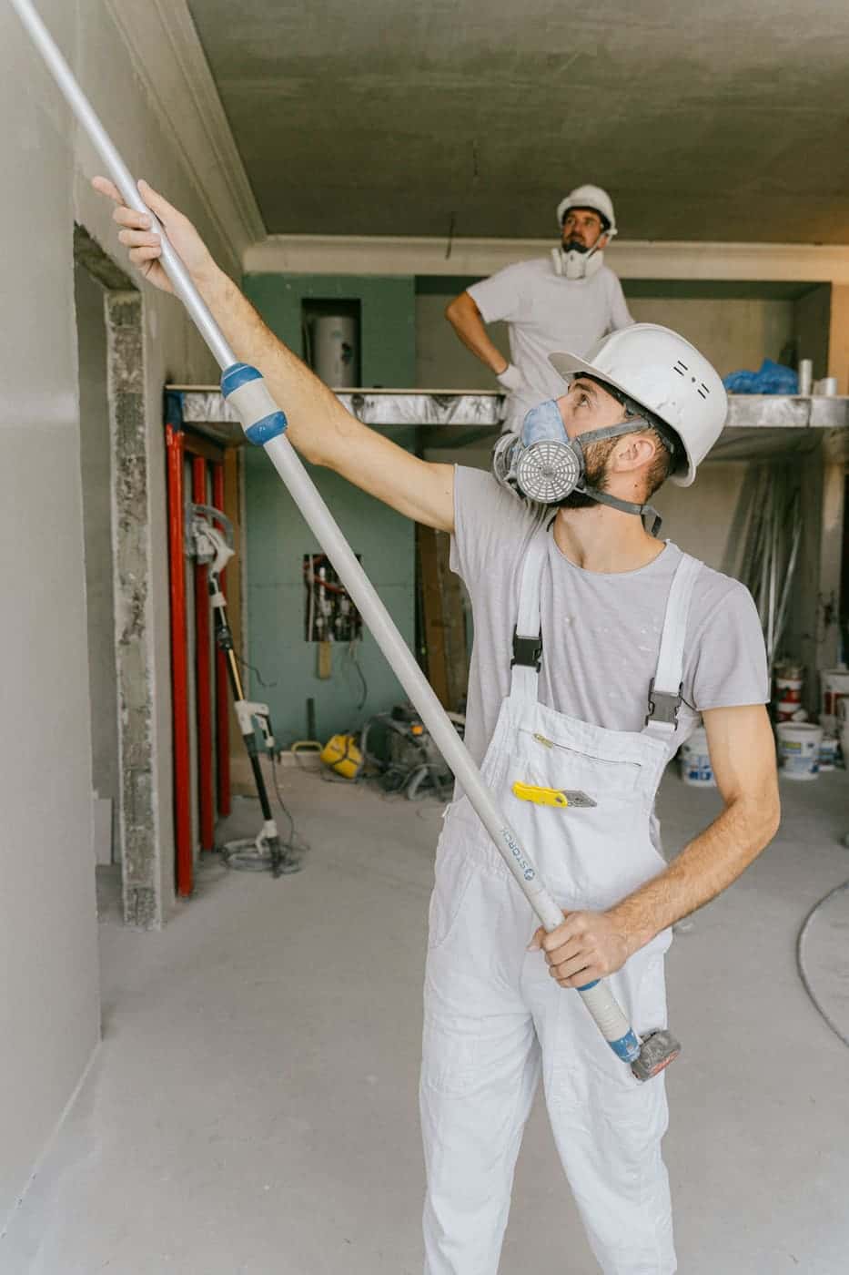 A worker in a respirator, hard hat and overalls works on the walls of a stripped-out room